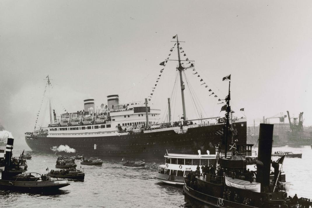 Black-and-white photograph of a large ship in the water with flags on two masts and covering lines from mast to deck. Smoke is billowing from the ship’s two smokestacks.