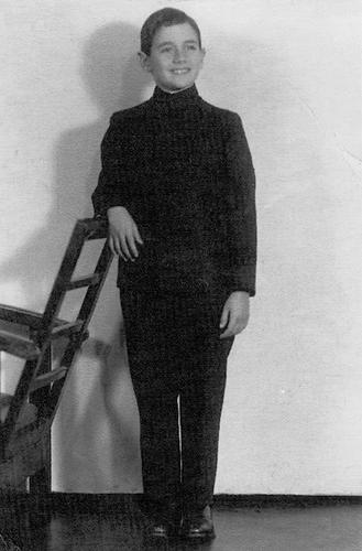 Un garçon vêtu d’une chemise et d’un pantalon noirs pose fièrement en regardant au loin. Son bras droit est posé sur le dossier d’une chaise.