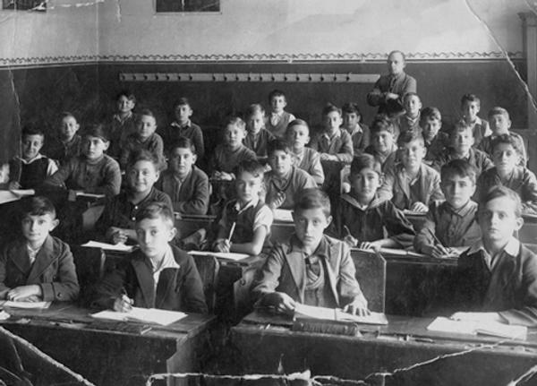 Classroom full of young boys sitting at desks with books open in front of them, looking at the camera, their teacher standing at the back of the classroom with his arms folded.