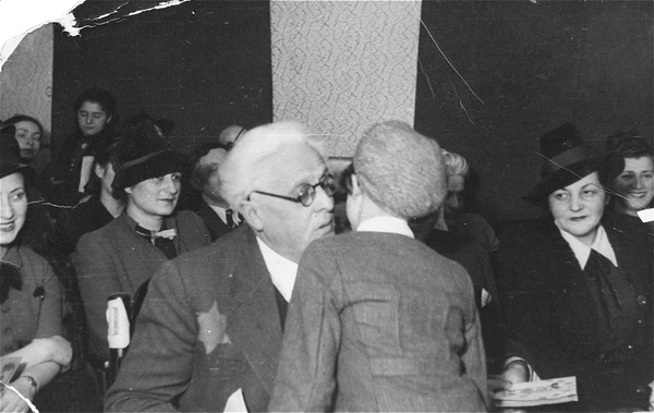 Un homme âgé portant une étoile à six branches embrasse un enfant sur la joue. Des femmes du public les observent avec tendresse.