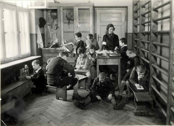 Dans une petite salle de gymnastique, des élèves s’occupent à des jeux. Une femme est debout derrière un pupitre.