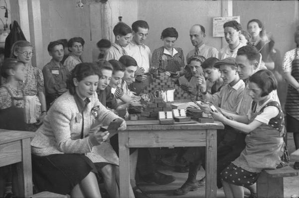 Debout ou assis, de jeunes adultes observent et manipulent les objets posés sur une table. Le chiffre 280 a été tracé en blanc dans le cadre du cliché.