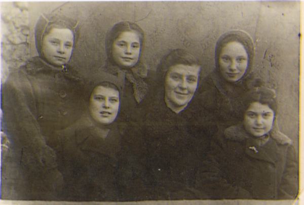 Cinq fillettes vêtus de chapeaux et de manteaux posent avec une femme. Elles affichent de timides sourires.