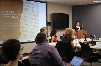 A woman speaks at a podium to a panel seated around a conference table. A projected slide reads Buried Words: A Workshop on Sexuality, Violence and Holocaust Testimonies.