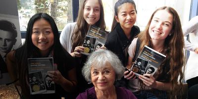 Four smiling young women hold up books while standing behind an older woman with gray hair seated in front of them. They are indoors, with light coming from large windows in the background.