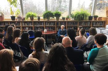 An older man speaks to a group of students seated in a library, with bookshelves and potted plants behind him and large windows letting in natural light.