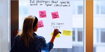 A woman writes on a large paper posted on a window, surrounded by colorful sticky notes with classroom strategies. The view outside shows blurred windows of another building.