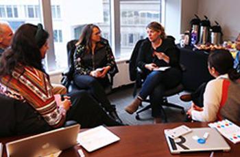 Five people sit in a small office, engaged in discussion. Papers, a laptop, and markers are on the table. Large windows show another building outside. Coffee dispensers and snacks are on a side table in the background.