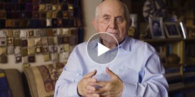 An older man in a light blue shirt sits indoors, gesturing with his hands. Behind him are shelves with books, framed photos, and colorful fabric squares on the wall. A play button is overlayed on top.